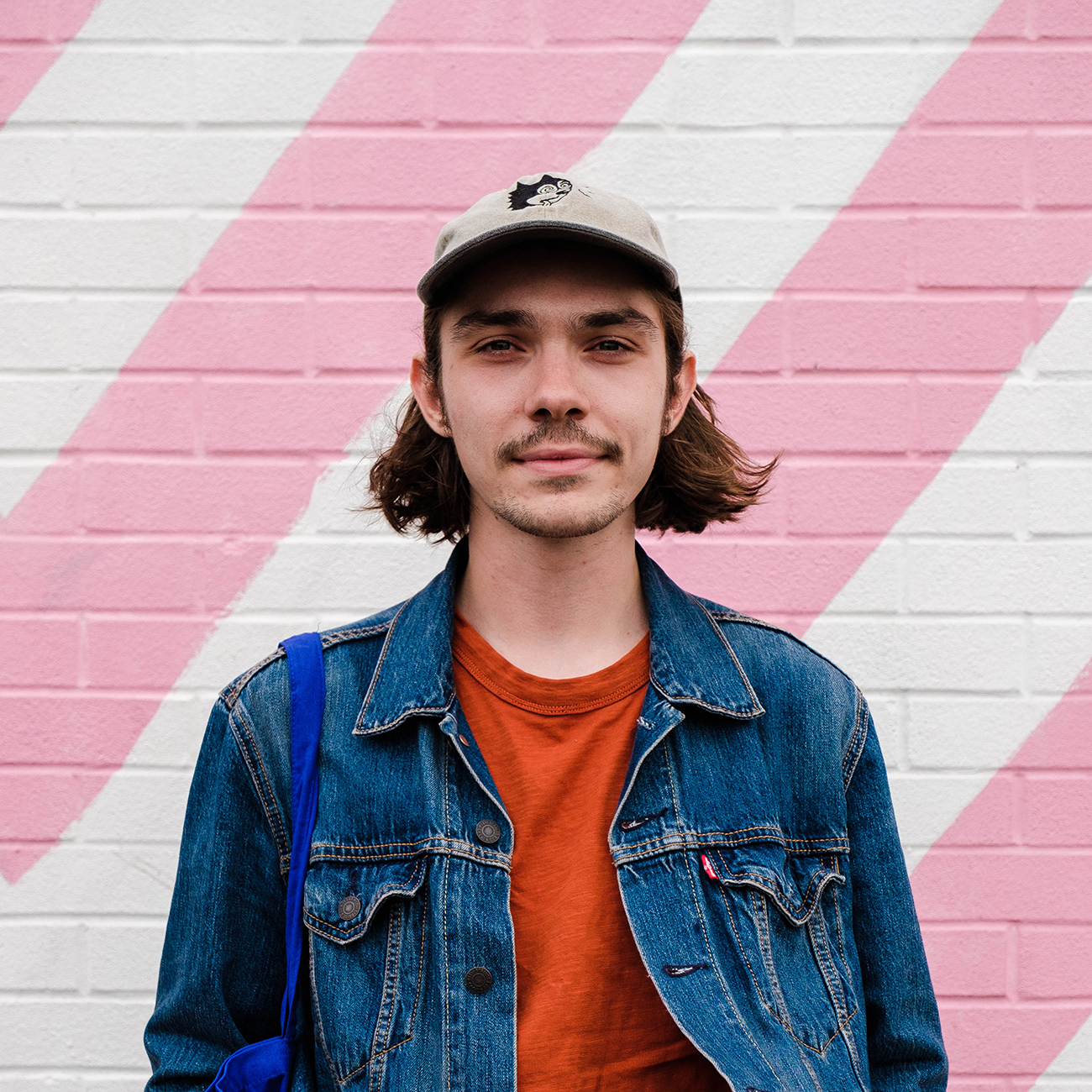 Young man in denim jacket and cap stands against a white and pink diagonal striped wall, carrying a blue bag.