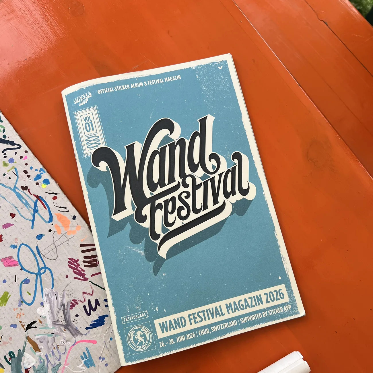 Wand Festival 2026 official sticker album and festival magazine, supported by StickerApp, next to a paint marker on an orange table