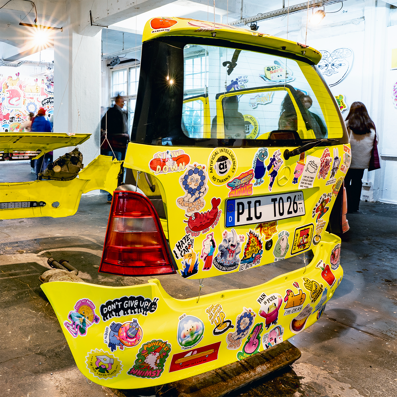 Yellow Mercedes body covered in bumper stickers in various shapes and sizes, with a german licence plate in a art exihibition context..