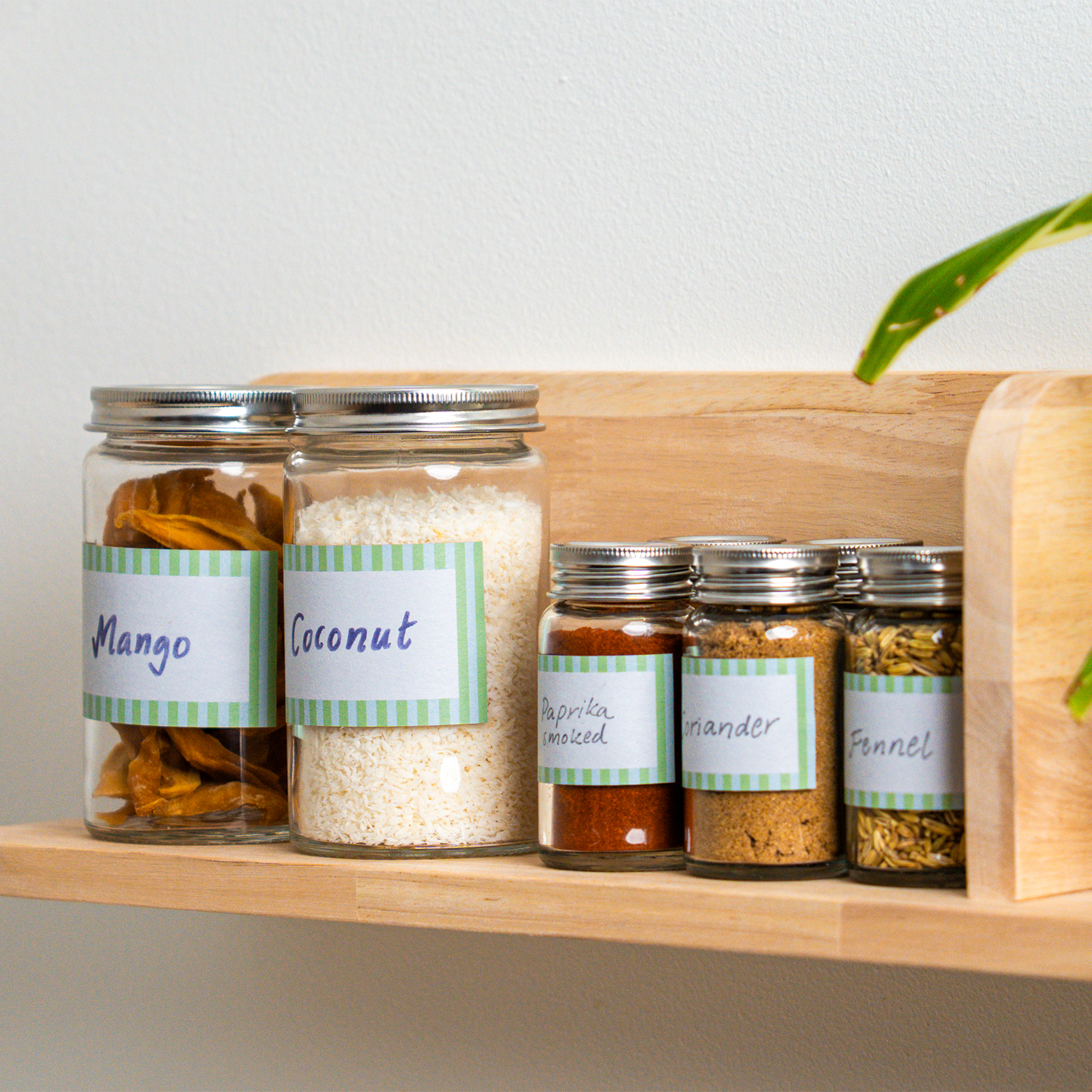 Glass jars with handwritten labels placed on a wooden kitchen shelf. The jars contain dried mango, coconut, and various spices including smoked paprika, coriander, and fennel seeds.