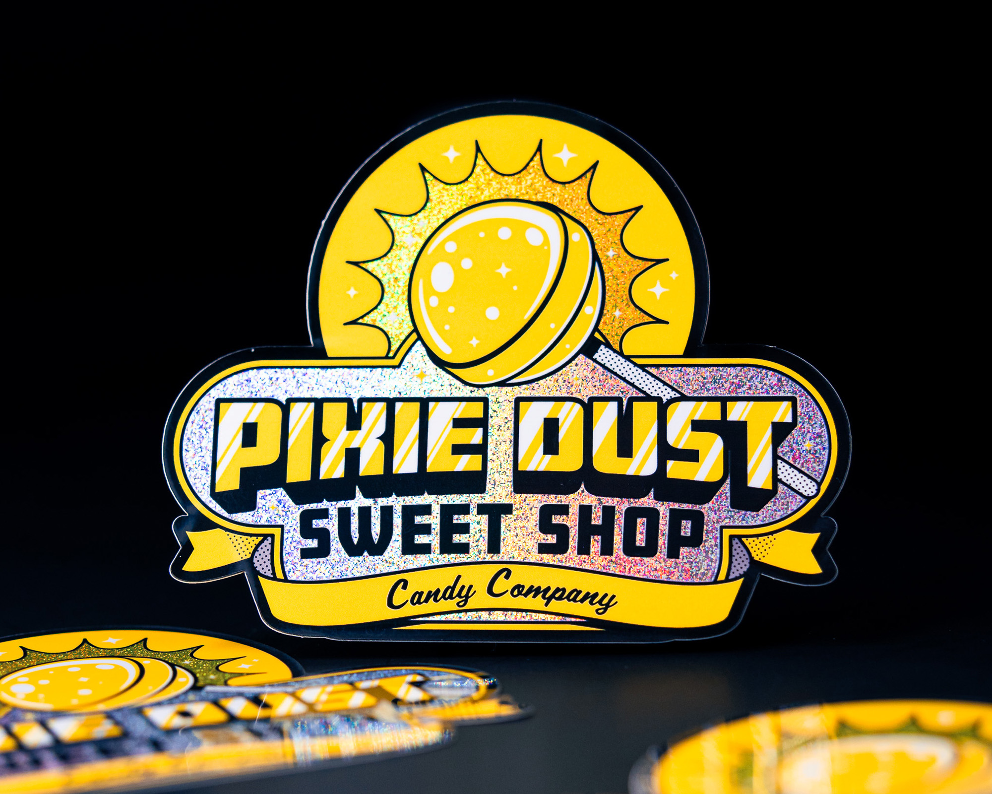 Yellow and glittery Pixie Dust Sweet Shop sticker featuring a lollipop design on a dark background.