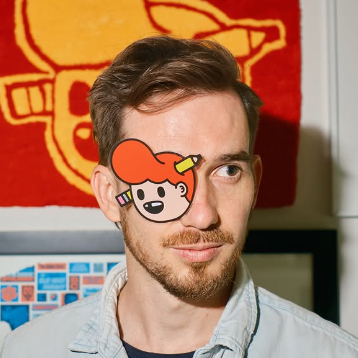 Tyler Pate aka The Creative Pain with a cartoon face sticker over one eye, standing in front of colorful artwork.